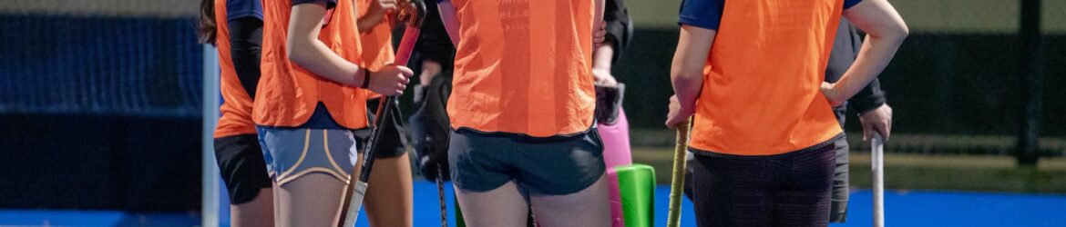 Hockey players in orange bibs listening to a coach during a training session